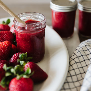 glass mason jar filled with strawberry honey butter with a wooden spoon sticking out of it on a white oval platter surrounded by fresh strawberries in the foreground and two closed jars of strawberry honey butter in the background.